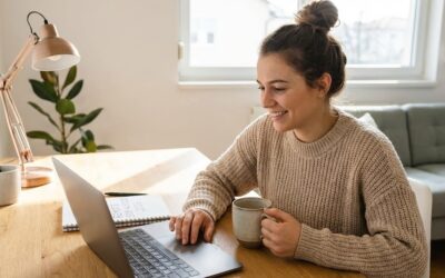 A smiling woman in a knitted sweater sitting at a home desk, working on a laptop and holding a mug, with a notebook beside her containing the handwritten text "REMOTE SIDE HUSTLES 2026".