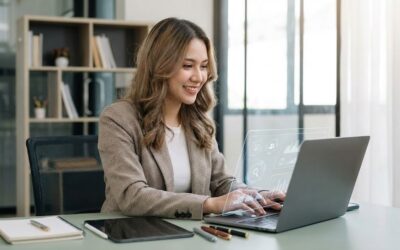 A smiling young woman in a plaid blazer sits at a desk in a modern office, typing on a laptop. A futuristic holographic display showing data charts and icons projects from the laptop's keyboard. Beside the laptop are a tablet, notebook, and pens. A bookshelf is visible in the background near a large window.